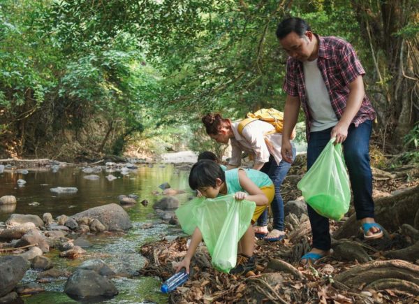 A family picks up trash together at the side of a small stream surrounded by trees.